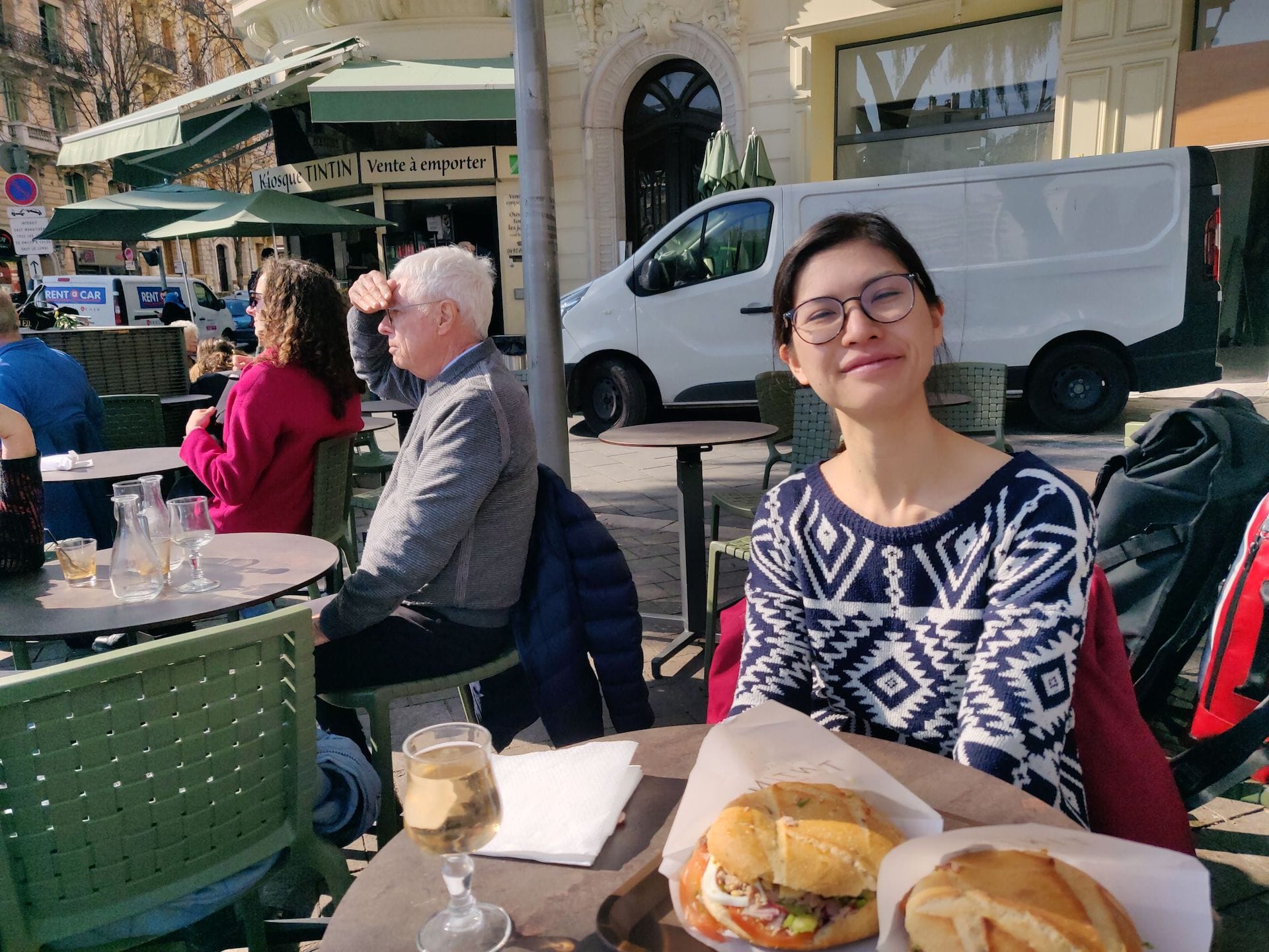 Clara à table sur la terrasse du Kiosque Tintin, qu'on aperçoit à l'arrière de la photo. Sur la table, deux pan bagnat et un verre de rosé.