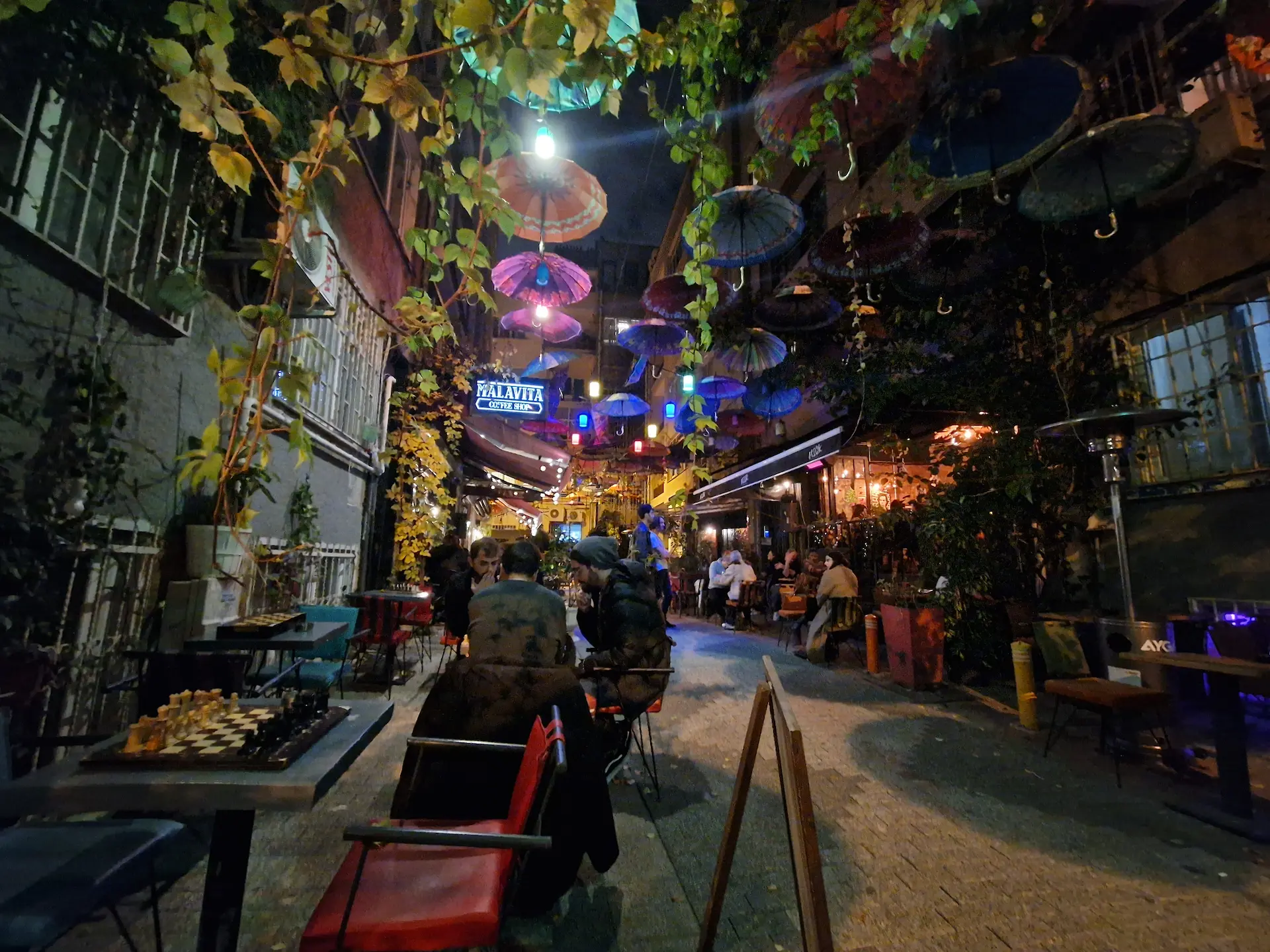 Une ruelle du quartier des bars de Kadıköy le soir, on voit des tables où sont placés des plateaux d'échecs, la ruelle est décorée de plantes grimpantes et de parapluies suspendus.