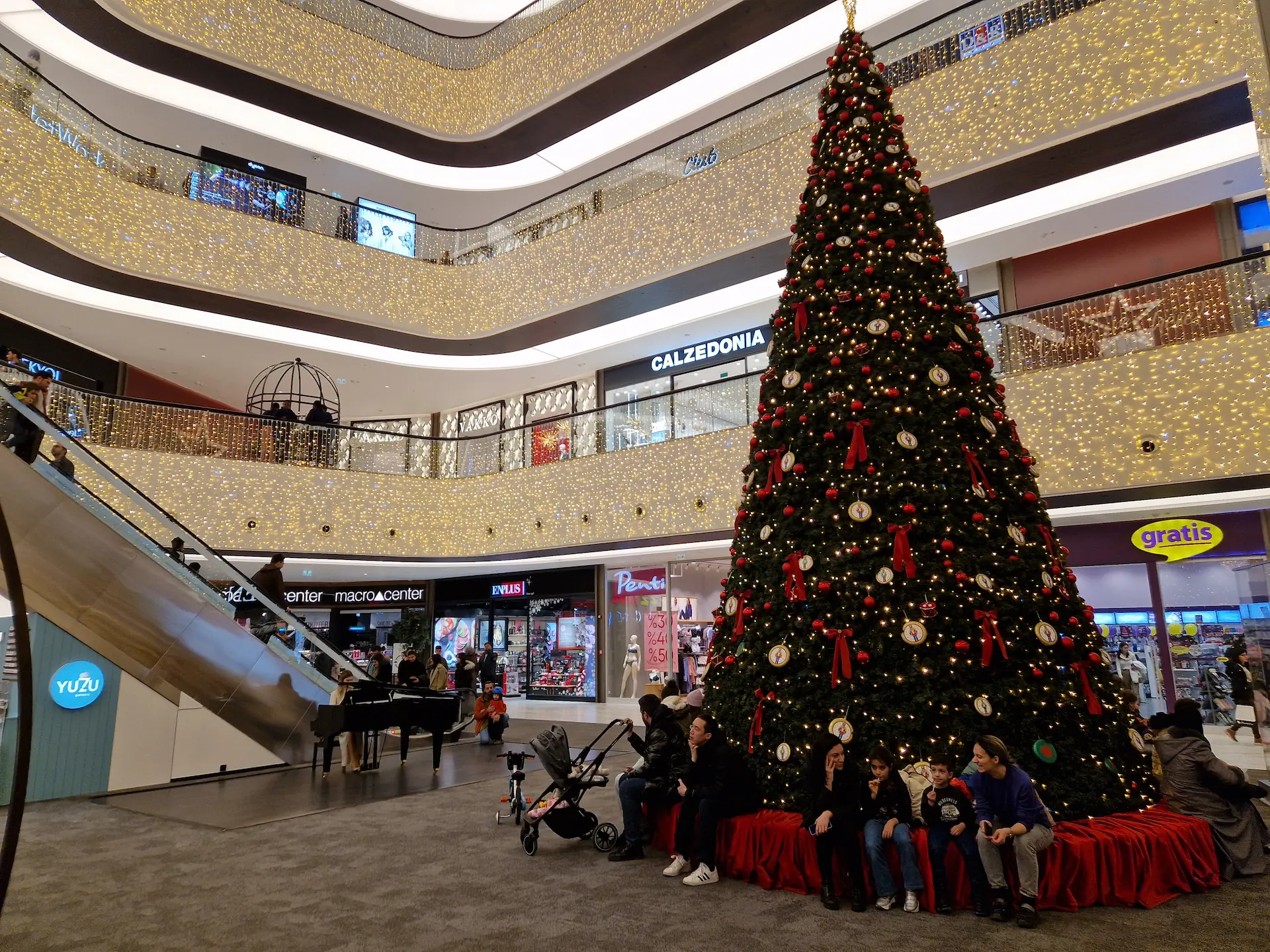 Dans le centre commercial : la pianiste est sur la gauche, le sapin illuminé est sur la droite. Des visiteurs sont assis au pied du sapin.