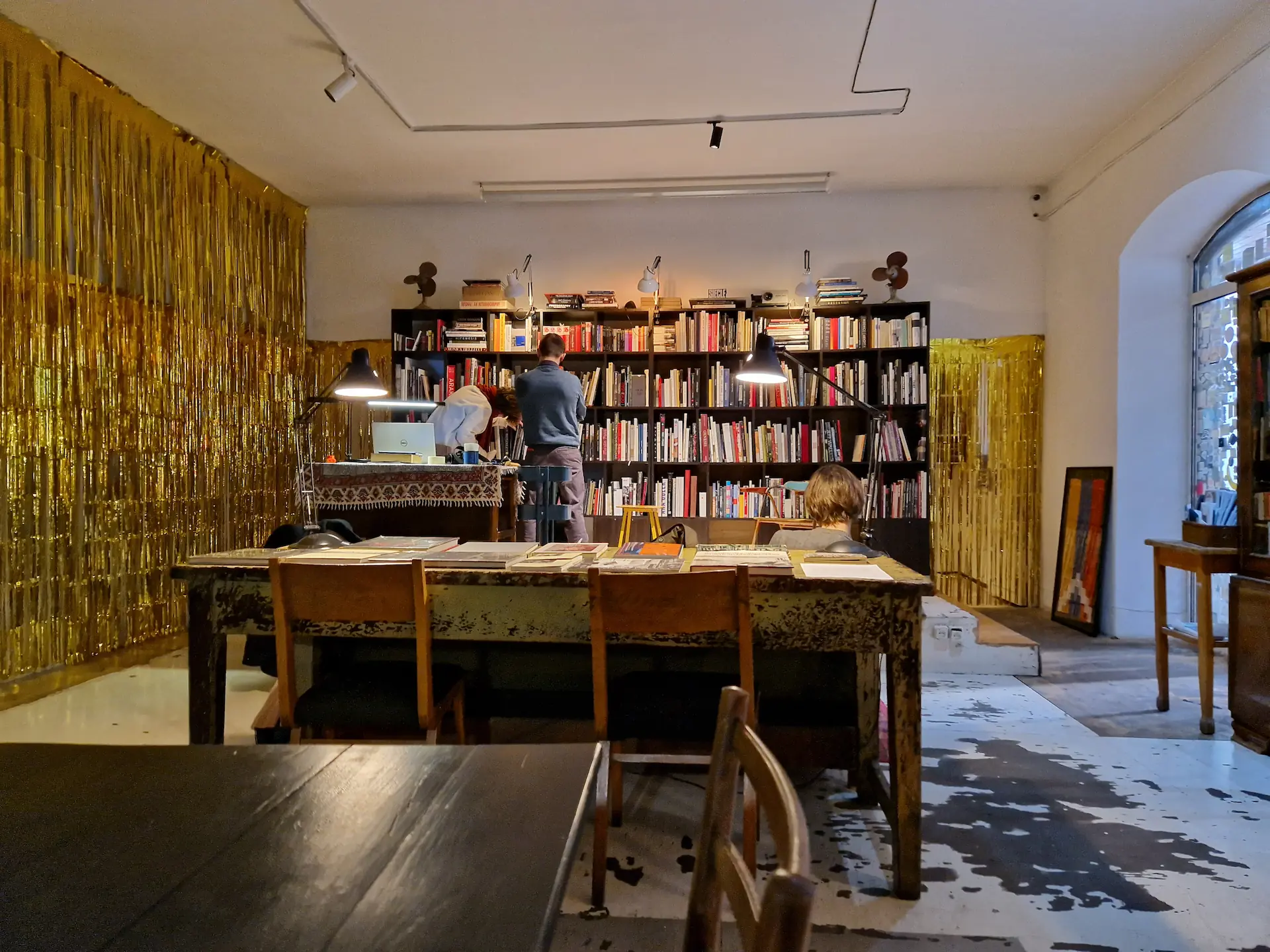 Dans le café-librairie Mirzoyan, on voit une bibliothèque avec des livres au fond de la pièce. Un canapé au centre de la pièce et des tables au premier plan.