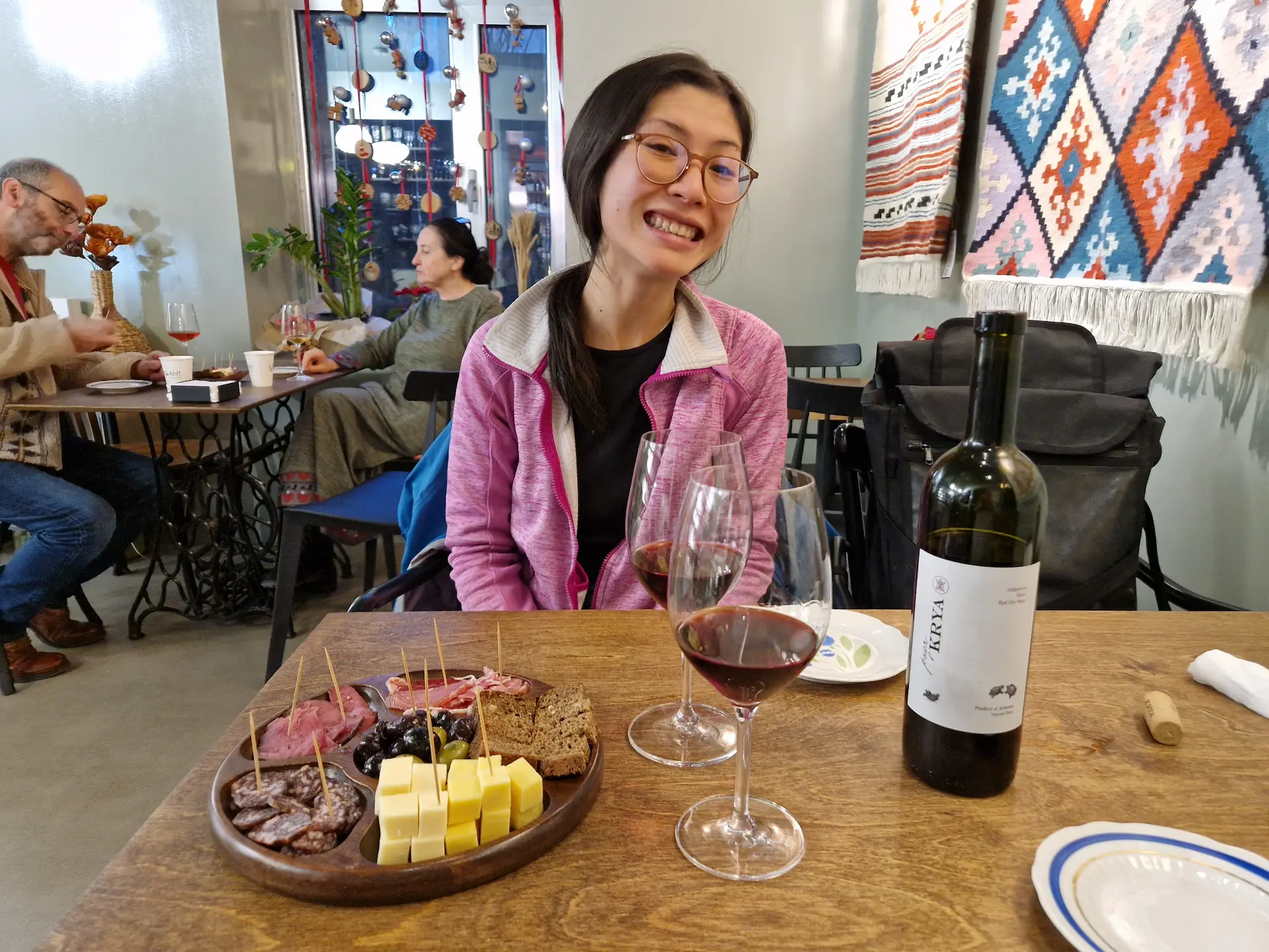 À une table du bar à vin, on déguste un verre de vin rouge arménien avec une planche de charcuteries et fromages locaux. Clara fait un grand sourire (légèrement forcé) à la caméra.