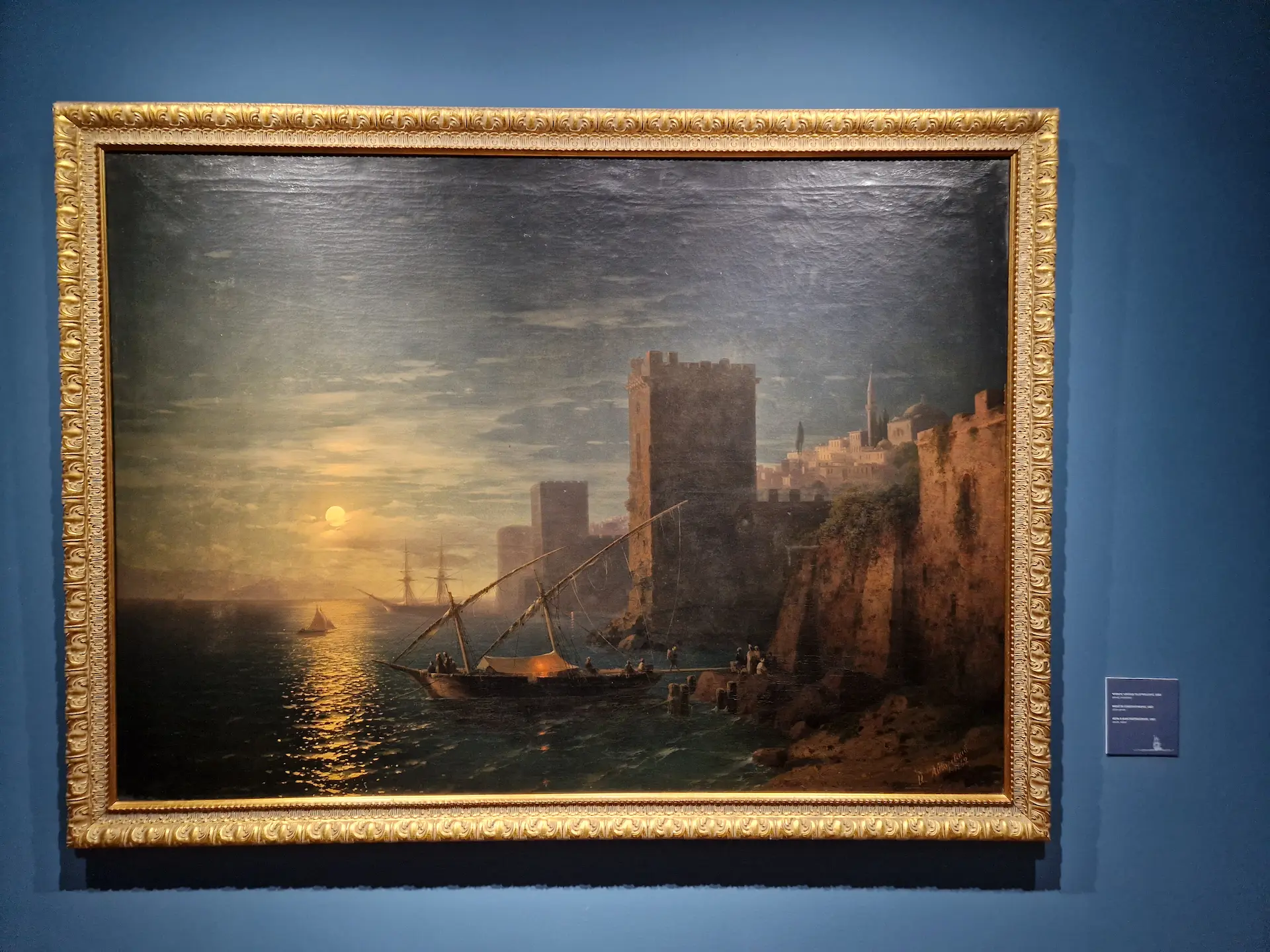 Peinture réaliste d'un coucher de soleil sur la côte, le soleil couchant se reflète dans la mer calme, les ramparts d'une ville s'élèvent sur la terre ferme, il y a un bateau de pêche au premier plan.