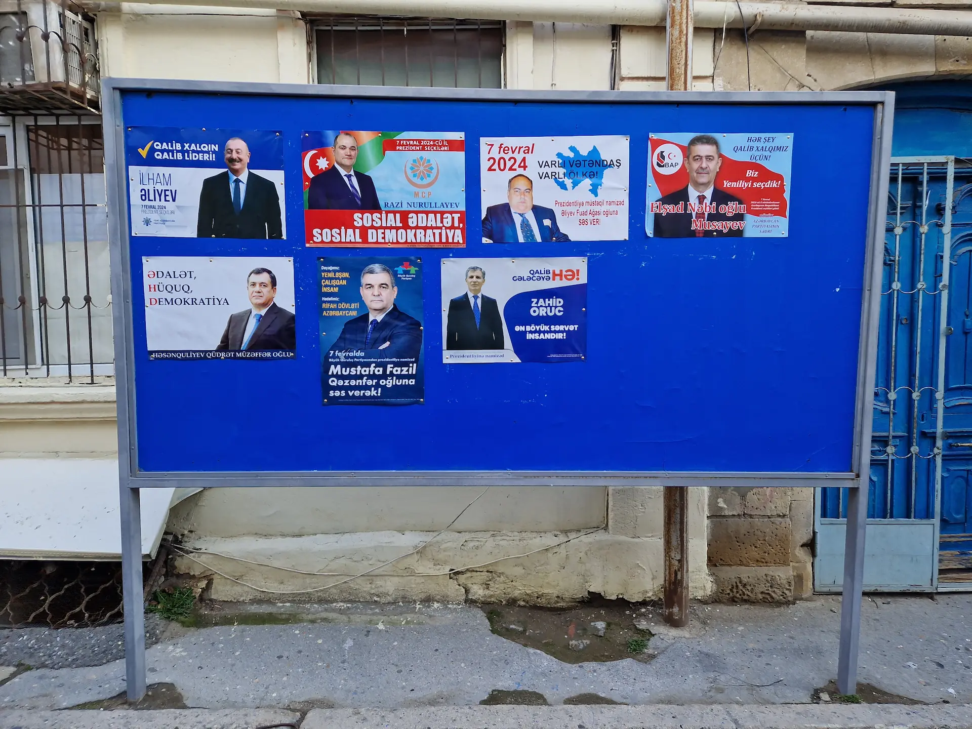 Un grand panneau bleu où sont accrochés les affiches des candidats. On en compte sept.