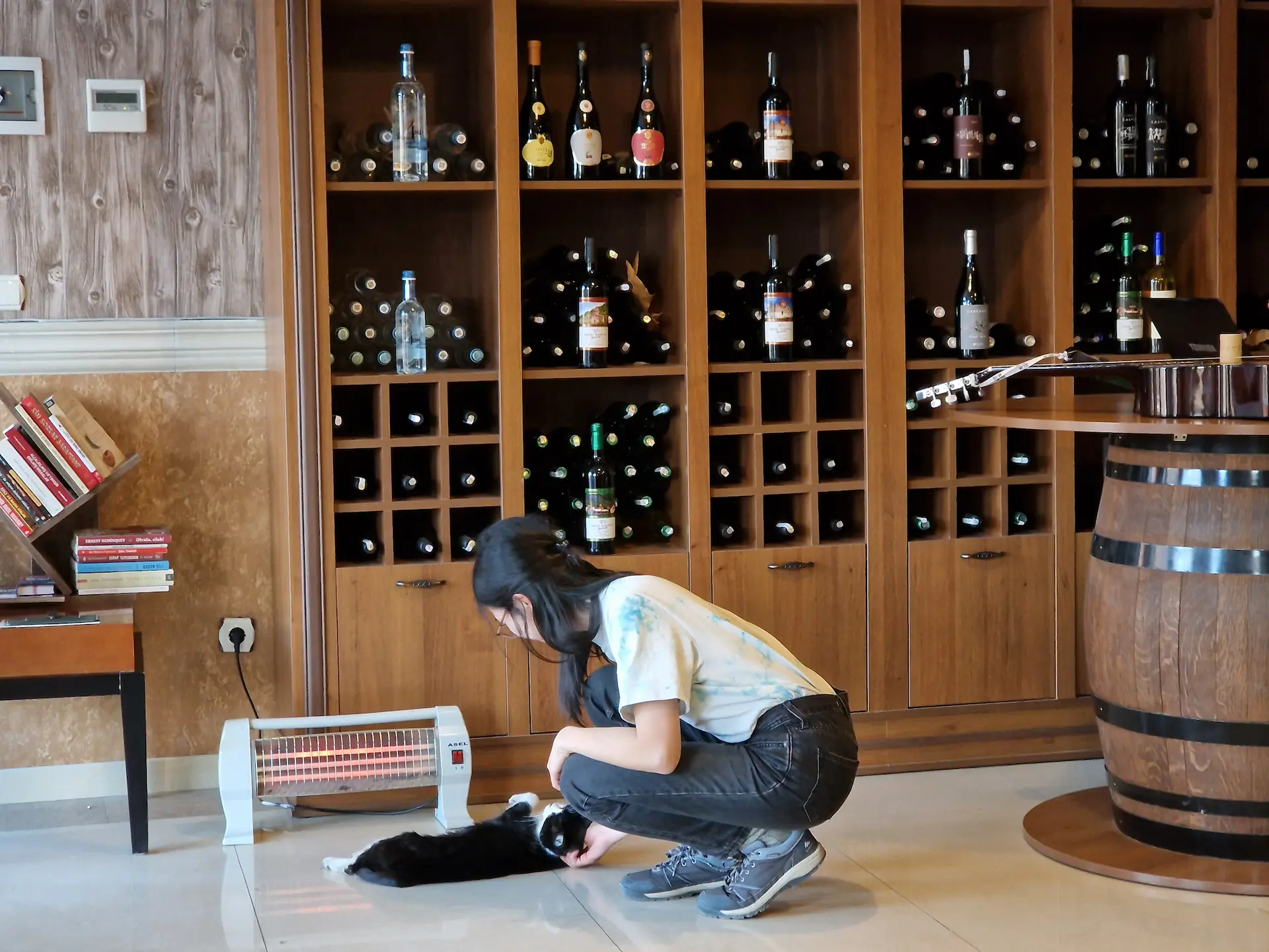 Clara fait des câlins au chat de la maison qui s'est couché juste devant le chauffage portable. Des étagères de vin sont en fond.