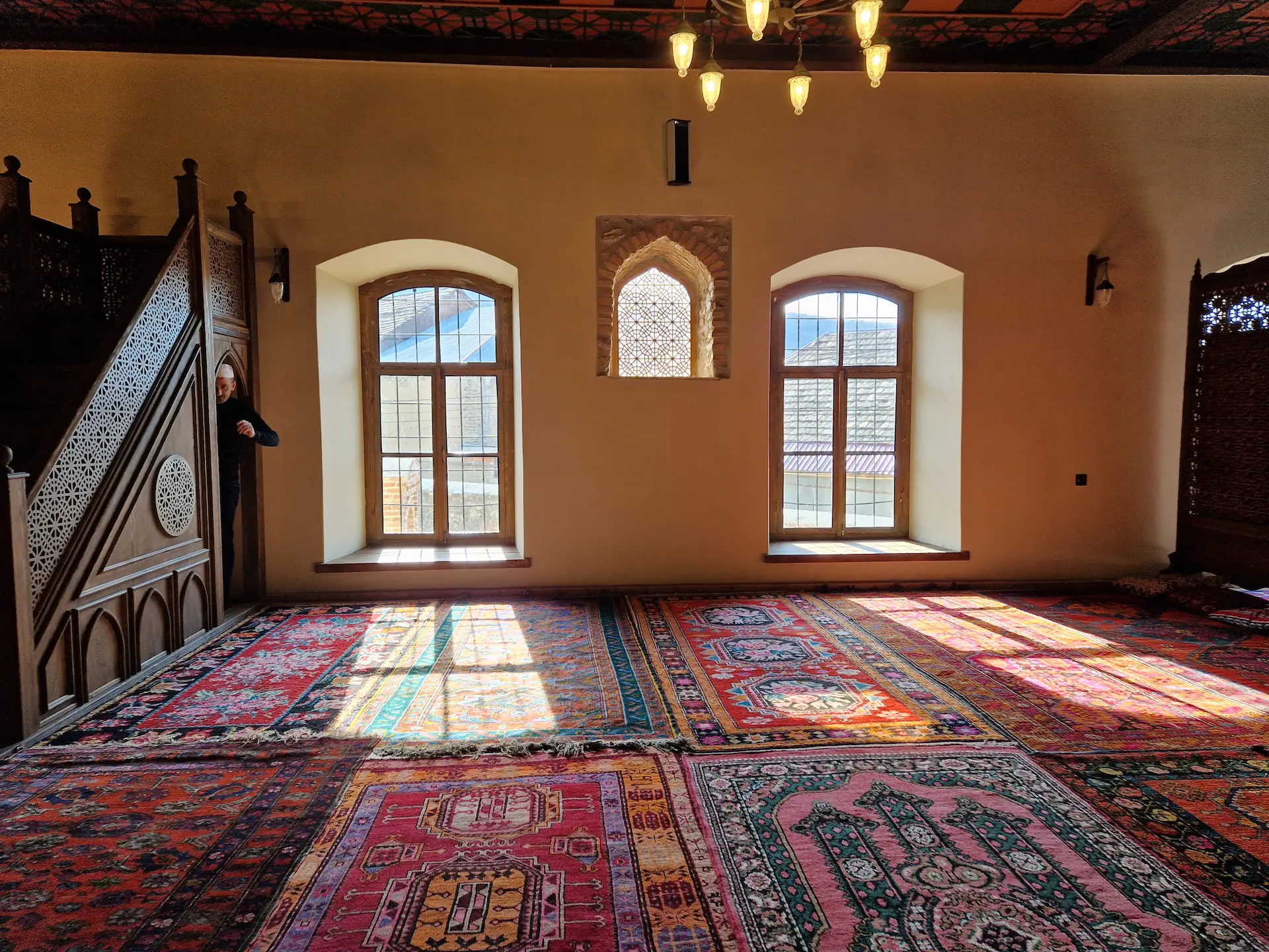 L'intérieur de la mosquée. Le sol est recouvert de tapis, on voit l'imam qui se prépare pour l'appel à la prière.