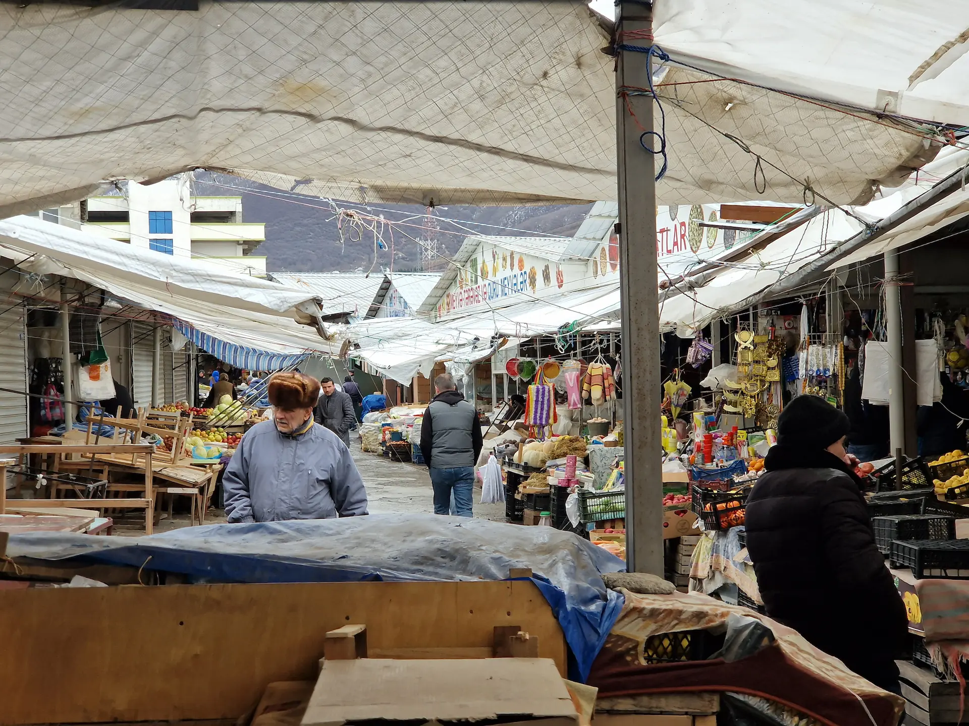 Une ruelle du marché, des vendeurs de légumes sur le côté. On voit un monsieur avec une grosse veste et un chapeau en fourrure marcher dans notre direction.
