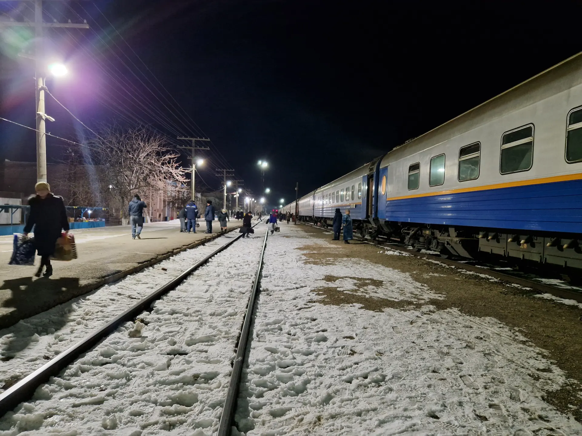 Il fait nuit, les gens traversent à pied des rails pour accéder à la voie où le train se trouve. Le sol et les rails sont couverts de neige. La lumière des lampadaires blanche éclaire fortement la gare.