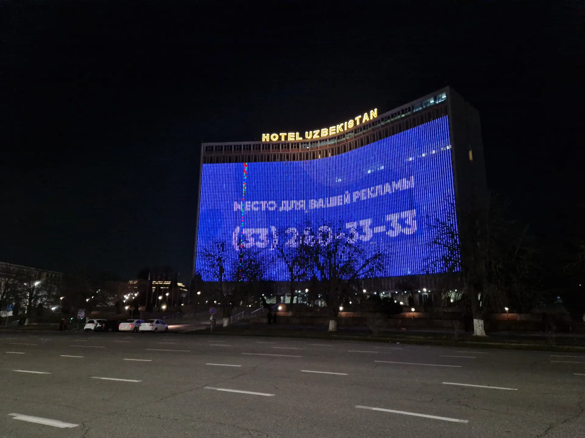L'hôtel Uzbekistan de nuit, les motifs de la façade ne sont pas visibles derrière l'affichage en LED d'un numéro de téléphone sur fond bleu