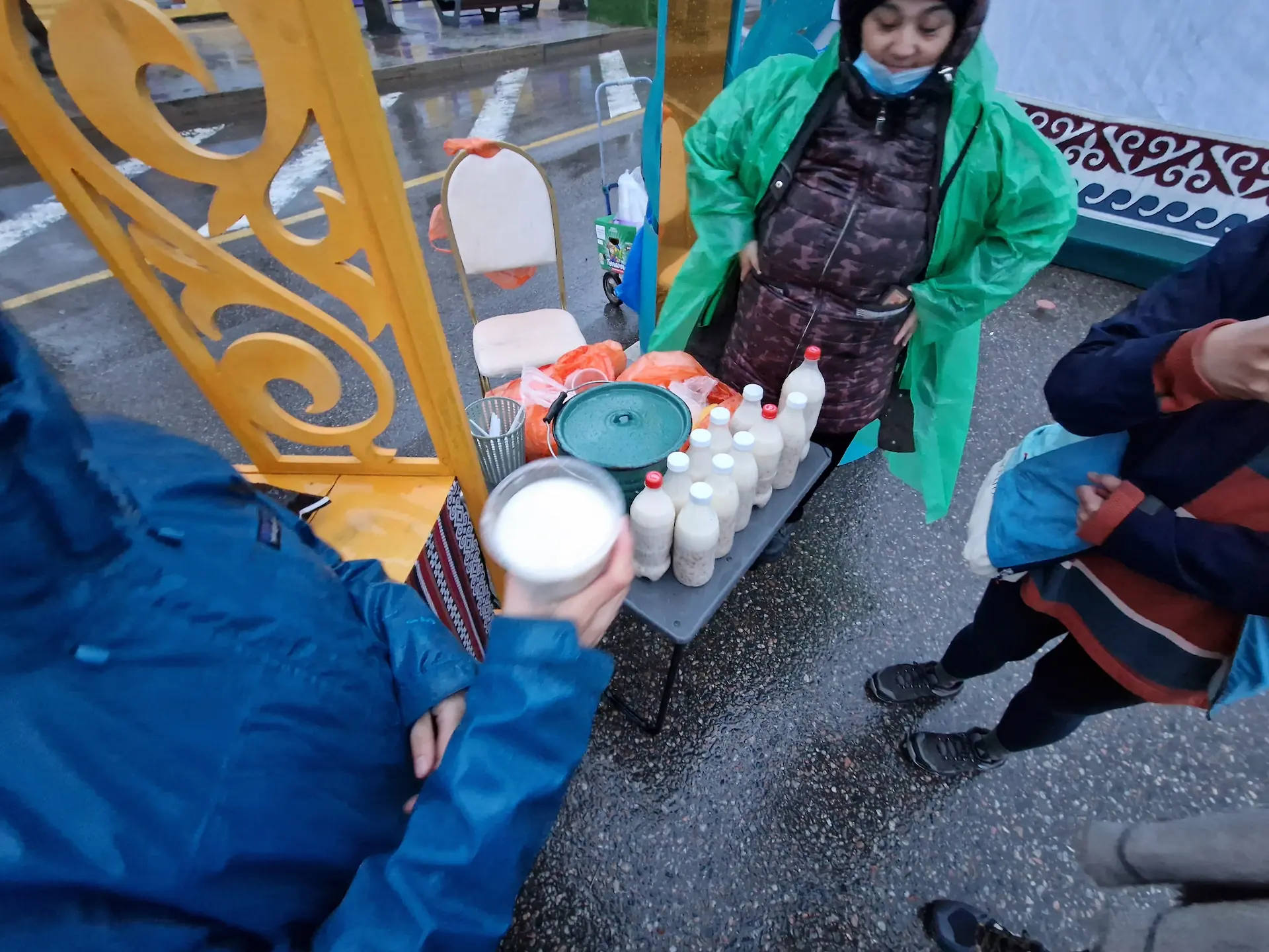 Clara achète un verre en plastique de Nauryz kozhe devant un patit stand où une dame le sert de bouteilles en plastique